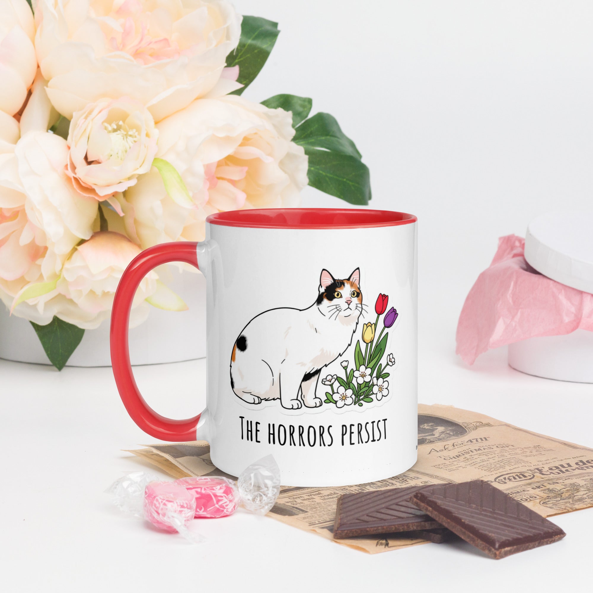 White ceramic mug with red rim and handle featuring calico cat Lina Love standing beside flowers and the text “The horrors persist, but so do the snacks.”