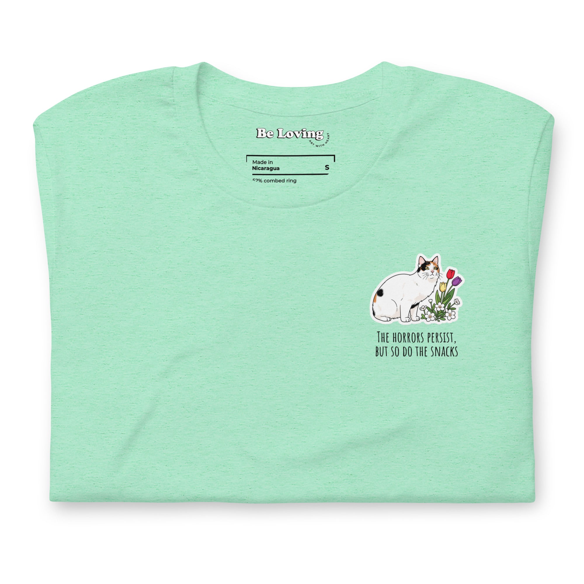 Mint green unisex t-shirt with Be Loving neck label and small Lina Love cat illustration with flowers and the text “The horrors persist, but so do the snacks” printed on the chest.